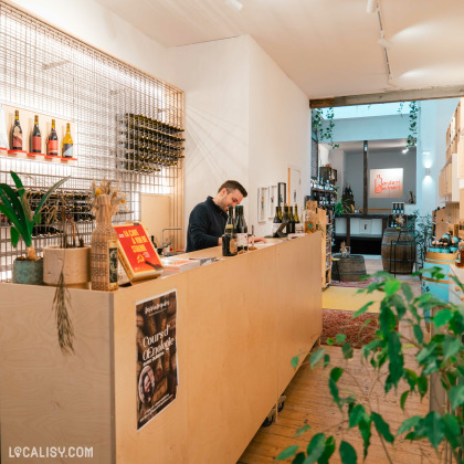 Un comptoir en bois avec une personne derrière, des étagères remplies de bouteilles, et des matériaux promotionnels chez “Les Vintrépides” à Liège.