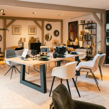 Une table à manger dressée avec des assiettes et des tasses, entourée de chaises, ainsi que divers objets décoratifs comme des œuvres murales, des miroirs et des étagères avec des objets au magasin “Home Design” à Neupré.