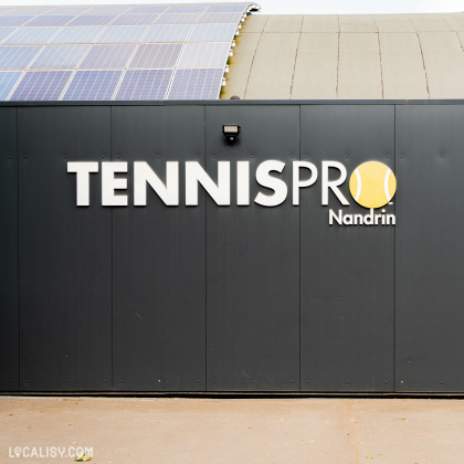 Devanture du magasin “Tennispro Nandrin” à Nandrin. Le mur est sombre avec le nom du magasin écrit en grandes lettres blanches. Au-dessus, un logo jaune représentant une balle de tennis est visible. Des panneaux solaires sont installés sur le toit du bâtiment.