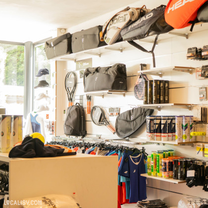 Des raquettes de tennis accrochées au mur, des vêtements sur des portants, et des étagères remplies de boîtes de balles de tennis et d’autres équipements chez “Tennispro Nandrin” à Nandrin.