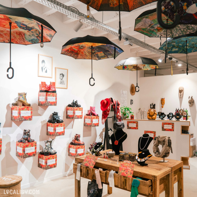 L’intérieur de la boutique “100% femme.bis” à Liège est décoré de parapluies colorés suspendus à l’envers au plafond, ajoutant une touche artistique unique à l’espace. Les murs sont ornés de divers articles, notamment des sacs à main sur des étagères et des bijoux exposés sur des présentoirs.