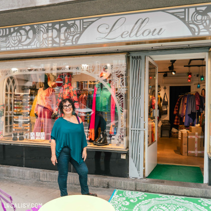 Devanture de la boutique “LELLOU” à Liège. La boutique a un auvent gris avec le nom du magasin en lettres cursives blanches et des motifs décoratifs. L’entrée est ouverte, révélant des vêtements colorés exposés. Une personne se tient devant la boutique, habillée de manière décontractée.