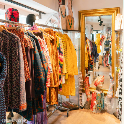 Des portants de vêtements colorés avec divers motifs, y compris des robes et des manteaux. Des accessoires comme des chapeaux et des sacs sont également visibles à l’intérieur de la boutique “LELLOU” à Liège.