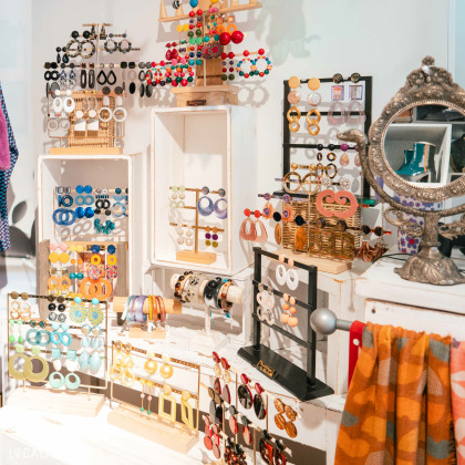 Plusieurs présentoirs avec une variété de lunettes colorées et de lunettes de soleil dans la boutique “LELLOU” à Liège. À gauche, une étagère blanche est remplie de lunettes, et au-dessus, un présentoir décoratif en forme d’arbre, également portant des lunettes.