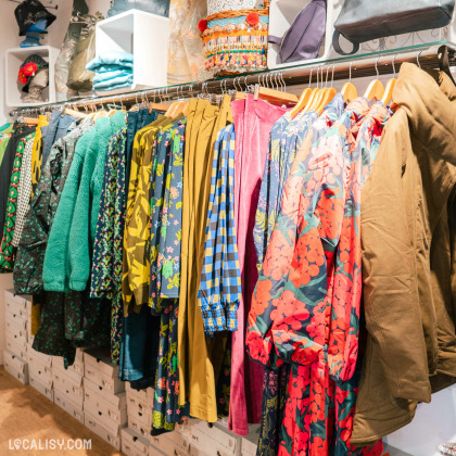 Une variété de vêtements colorés suspendus à un portant, avec des motifs allant du floral au géométrique dans la boutique “LELLOU” à Liège. Les vêtements sont principalement des vestes et des chemises.