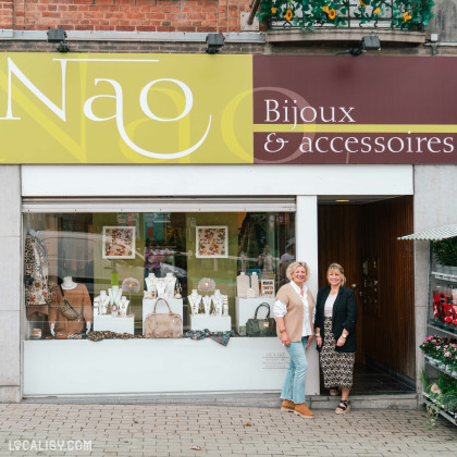 Devanture du magasin “Nao” à Liège, spécialisée dans les bijoux et accessoires. La vitrine expose divers articles, avec une enseigne verte et jaune au-dessus de l’entrée. Deux personnes se tiennent devant le magasin.