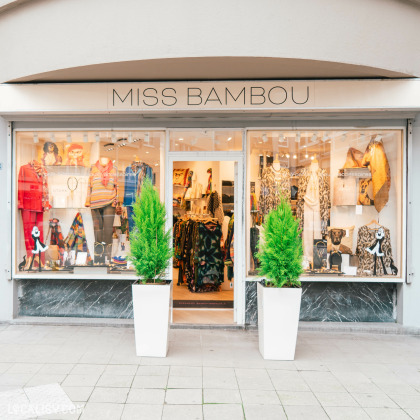 Devanture du magasin “Miss Bambou” à Liège. La façade est courbée avec le nom du magasin affiché en haut. Les vitrines de chaque côté de la porte présentent divers vêtements et accessoires, avec des mannequins habillés de tenues colorées.