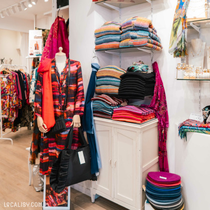 Des vêtements colorés et des accessoires, avec des motifs variés. Des textiles pliés sont disposés sur des étagères et un mannequin porte un vêtement vibrant orange et bleu au magasin “Miss Bambou” à Liège.