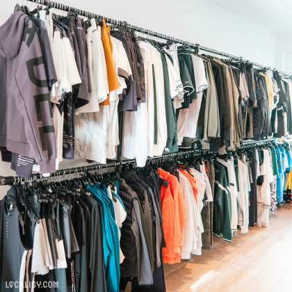 L’intérieur du magasin “NuevoPadel” à Tilff, avec des vêtements suspendus sur des portants, principalement des t-shirts et des hauts, organisés par couleur.