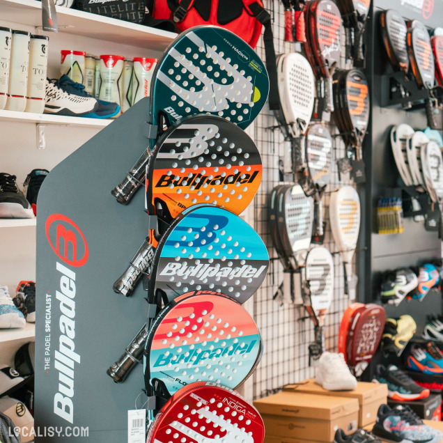 Un mur exposant divers raquettes de padel et des boîtes de chaussures en dessous au magasin “NuevoPadel” à Tilff. La marque “Bullpadel” est bien visible, avec des raquettes de différentes couleurs comme l’orange, le bleu et le rose.