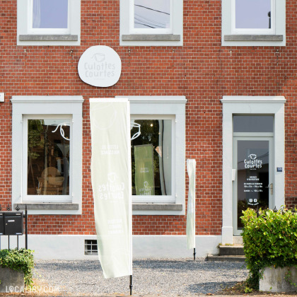 Devanture du magasin “Culottes Courtes” à Heusy, avec une façade en briques et deux grandes vitrines exposant des articles. Un panneau circulaire blanc avec le nom du magasin est visible au-dessus de la vitrine de gauche.