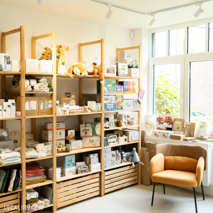 Des étagères en bois bien organisées exposant divers articles pour enfants, comme des livres, des jouets et des vêtements à l’intérieur du magasin “Culottes Courtes” à Heusy.