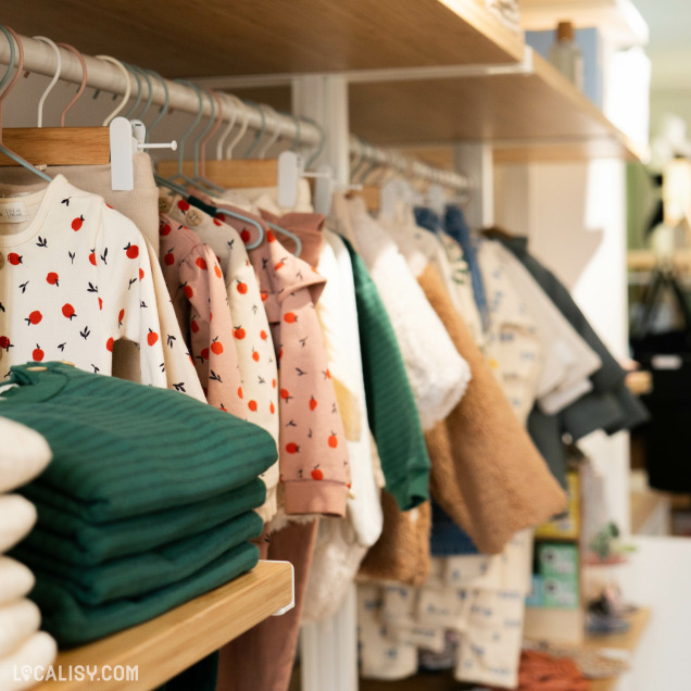 Divers vêtements pour enfants suspendus sur des cintres en bois et des étagères. Les vêtements sont colorés et variés, créant une ambiance accueillante et organisée au magasin “Culottes Courtes” à Heusy.