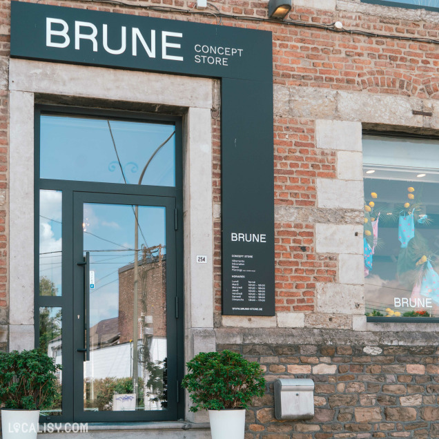 Devanture du magasin “BRUNE Concept Store” à Beaufays. La boutique a un auvent noir avec des lettres blanches indiquant “BRUNE CONCEPT STORE”. À droite de l’entrée, un tableau noir liste des catégories ou articles du magasin.