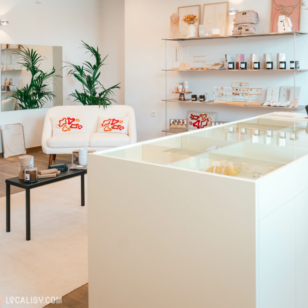 L’intérieur de “BRUNE Concept Store” à Beaufays avec des murs blancs et un sol en bois. Il y a deux fauteuils blancs avec des coussins décoratifs rouges, plusieurs étagères murales exposant divers articles, et une table centrale avec d’autres produits.