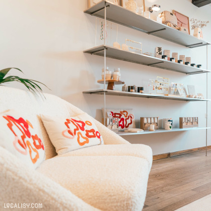 Un coin cosy du magasin “BRUNE Concept Store” à Beaufays, avec un canapé beige et des coussins rouges. En arrière-plan, des étagères murales présentent divers articles comme des bougies et des objets décoratifs.