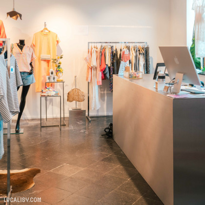 Des vêtements pour femmes sur des portants et des mannequins, un banc en bois en forme d’animal, et un comptoir de vente moderne avec un ordinateur à l’intérieur du magasin “BRUNE Concept Store” à Beaufays..