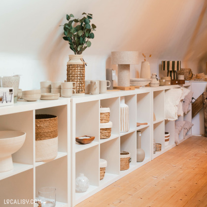 L’intérieur du magasin “Le Cottage Interior” à Charneux, avec une étagère blanche remplie d’articles de décoration comme des céramiques et des textiles.