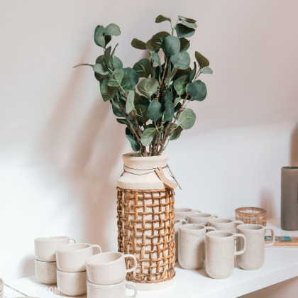 Etagère avec un vase en osier contenant des branches d’eucalyptus, accompagnée de tasses en céramique et de petits récipients. Ce présentoir du magasin “Le Cottage Interior” à Charneux met en avant un style de design intérieur rustique et naturel.