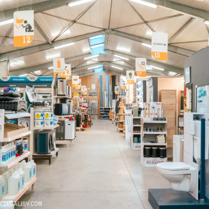 L’intérieur du magasin “Garsou” à Theux. On y voit une allée avec divers matériaux et produits de bricolage, tels que des toilettes, des éviers et des seaux de peinture.