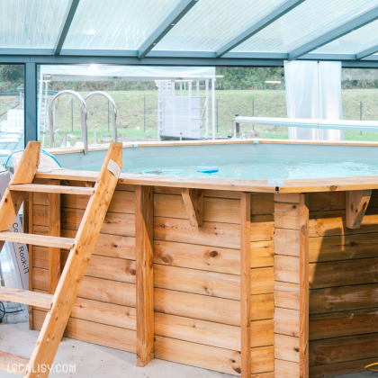 Une structure en bois à l’extérieur du magasin "Garsou" à Theux, peut-être un comptoir de bar ou de service, devant une piscine couverte par une bâche bleue. La structure est sous un abri avec un toit transparent.