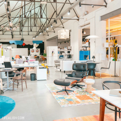 L'intérieur du magasin “Oburo” à Liège, avec un showroom spacieux présentant divers meubles de bureau. On y voit des bureaux, des chaises, des étagères et des luminaires sous un plafond haut avec des poutres métalliques apparentes.