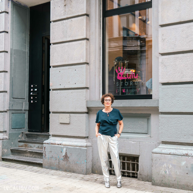 La devanture du magasin “Lulu Store” à Liège avec une grande fenêtre, le nom “Lulu” écrit en néon rose cursif, et en dessous, un texte plus petit indiquant “store”. Devant le magasin, une personne se tient debout, portant un haut bleu foncé et un pantalon clair.