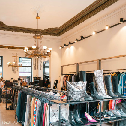 Divers vêtements et chaussures exposés sur des étagères et des présentoirs au magasin “Lulu Store” à Liège. Le magasin a un design élégant avec des lustres, des moulures décoratives au plafond et une ambiance bien éclairée qui met en valeur les articles.