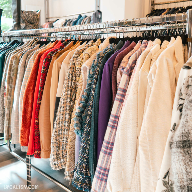 Divers vêtements suspendus sur un portant dans le magasin "Lulu Store" à Liège. Les vêtements incluent des pulls, des vestes et des chemises avec différents motifs et couleurs comme le rouge, le bleu, le violet et des designs à carreaux.