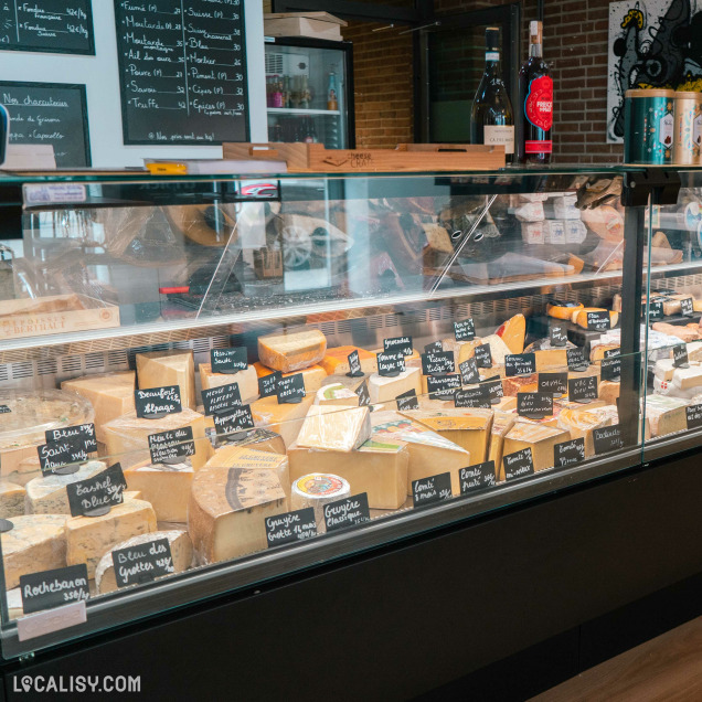 Un étalage de fromages à la fromagerie “L’affine Bouche” à Liège. Divers fromages sont soigneusement disposés derrière un comptoir en verre, chacun étiqueté avec de petits panneaux noirs.