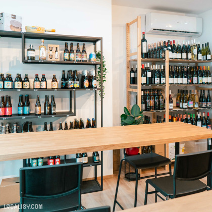 L’intérieur de la fromagerie “L’affine Bouche” à Liège, avec des étagères en bois remplies de bouteilles de vin et de bière, une longue table en bois avec des chaises noires autour, et quelques plantes vertes.