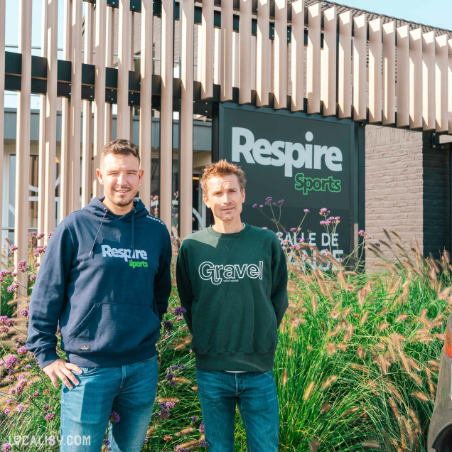 Devanture du magasin "Respire Sports" à Jalhay. Deux personnes debout devant le magasin, elles portent des sweats à capuche avec les mots “Respire” et “Gravel”. La façade est moderne avec des lattes en bois et de la végétation au premier plan.