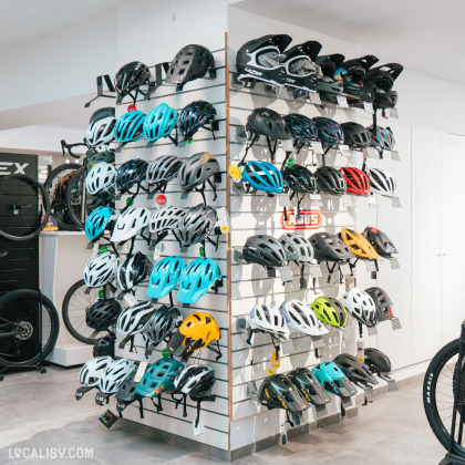 L’intérieur du magasin “Respire Sports Bike” à Spa, avec un présentoir central de casques de vélo de différentes couleurs et designs. À gauche, on aperçoit une partie d’un vélo, et à droite, une enseigne néon “SALE” en lettres rouges indique une promotion.