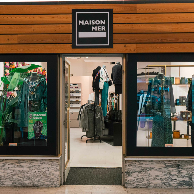 Devanture du magasin “Maison Mer” à Liège, avec une enseigne noire et des lettres blanches au-dessus de l’entrée. Le magasin a deux grandes vitrines de chaque côté de la porte centrale, exposant divers articles, notamment des vêtements et des plantes.