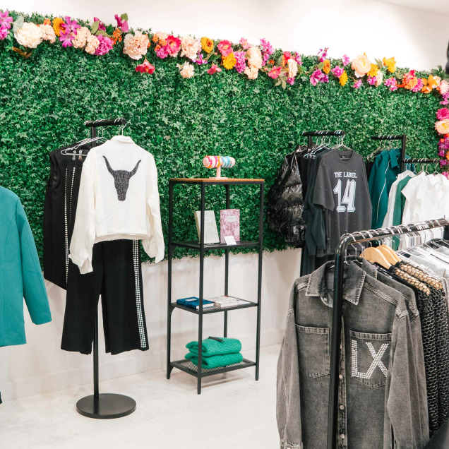 L’intérieur du magasin “Maison Mer” à Liège. On y voit un mur vert décoré de fleurs colorées, avec des vêtements exposés sur des cintres et des mannequins.