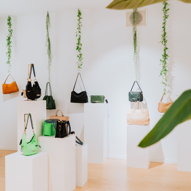 Des murs blancs et plusieurs socles blancs exposant une variété de sacs. Des plantes vertes suspendues décorent l’espace du magasin “Maison Mer” à Liège.