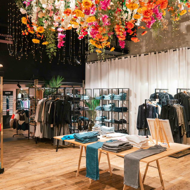 Le magasin “Maison Mer” à Liège présente un design moderne et élégant avec des fleurs colorées suspendues au plafond, créant un affichage vibrant. Les vêtements sont soigneusement disposés sur des étagères et des portants.