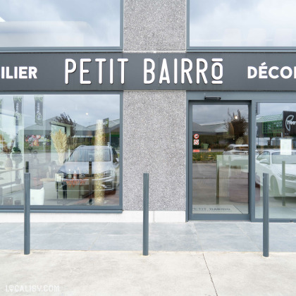 Devanture du magasin “Petit Bairro Déco” à Saint-Georges-sur-Meuse. La façade est grise avec de grandes fenêtres et une porte en verre. Le nom du magasin est écrit en lettres blanches audacieuses. Devant l’entrée, il y a deux bornes noires et une voiture est visible à travers l’une des fenêtres.