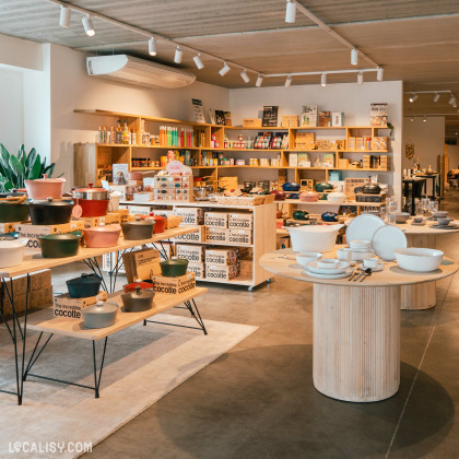 L’espace du magasin “Petit Bairro” à Saint-Georges-sur-Meuse est bien éclairé et présente une variété de produits exposés sur des tables et des étagères, y compris des livres, des ustensiles de cuisine et des plantes.