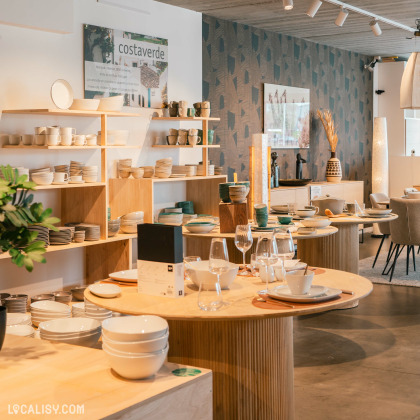 Une variété d’articles pour la maison et de vaisselle, soigneusement exposés sur des étagères en bois et des tables rondes à l’intérieur du magasin “Petit Bairro” à Saint-Georges-sur-Meuse.