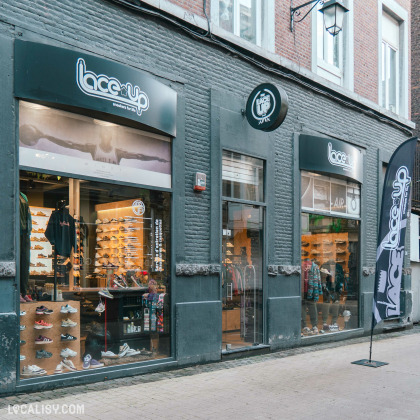 Devanture du magasin “Lace-Up” à Liège? la façade est gris foncé avec le nom et le logo du magasin au-dessus des grandes vitrines. Les vitrines exposent diverses chaussures et quelques vêtements. Un panneau sur le côté droit présente un branding supplémentaire pour “Lace-Up”.