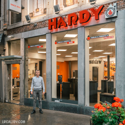 Devanture du magasin “Maison Hardy” à Liège. Le nom du magasin est affiché en grandes lettres rouges au-dessus de l’entrée, avec un logo comprenant un cercle rouge et un point blanc. La façade grise comporte de grandes vitrines qui permettent de voir l’intérieur bien éclairé du magasin, où divers articles sont exposés.