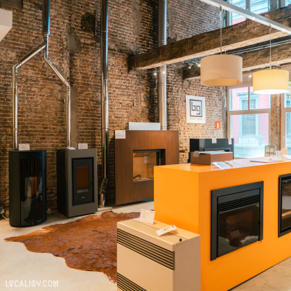 L’intérieur du magasin “Maison Hardy” à Liège, avec des murs en briques apparentes et un plafond haut de style industriel. Au premier plan, un comptoir orange vif avec des vitrines en verre expose divers modèles de poêles ou de chauffages.