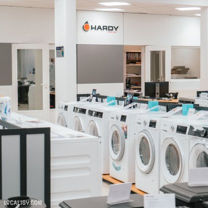 l’intérieur du magasin “Maison Hardy” à Liège, avec plusieurs machines à laver exposées au premier plan. En arrière-plan, d’autres appareils électroménagers sont visibles. Le logo du magasin, “HARDY”, est affiché sur un mur.