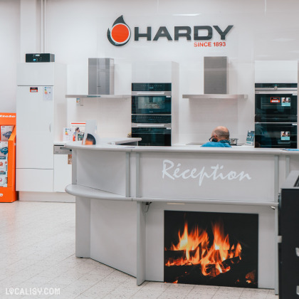 L’intérieur du magasin “Maison Hardy” à Liège, avec un comptoir de réception orange vif et des étagères remplies de divers articles en arrière-plan. Le design est moderne, avec des murs en briques apparentes et des luminaires suspendus.