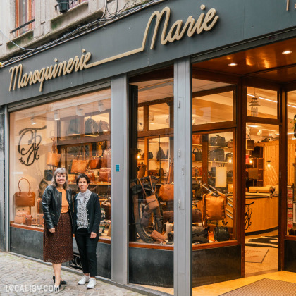 Devanture de la "Maroquinerie Marie" à Liège. Le magasin a un auvent noir avec le nom en lettres dorées. La vitrine expose divers articles en cuir. Deux personnes se tiennent devant le magasin, l’une partiellement cachée par l’autre.