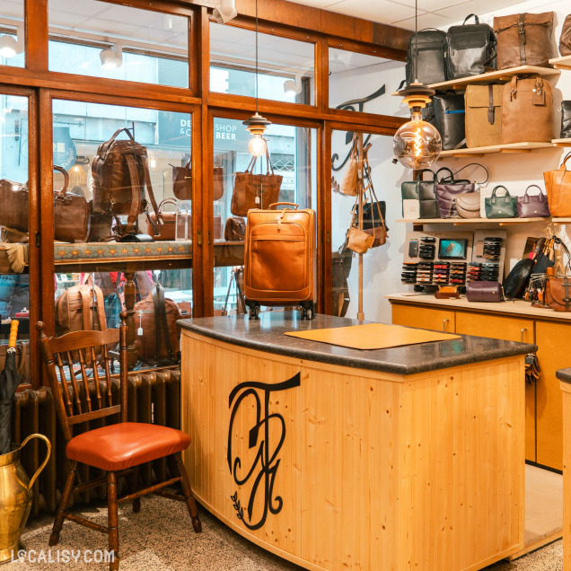 L’intérieur de la "Maroquinerie Marie" à Liège. On y voit divers articles en cuir, notamment des sacs et des accessoires, exposés sur des étagères et accrochés aux murs. Un comptoir en bois avec un logo orné “M” est au premier plan, et une chaise est placée à côté.