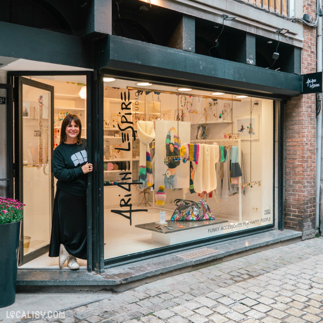 Devanture du magasin "Ariane Lespire" à Liège, avec une grande vitrine affichant divers articles colorés et accessoires. Le nom du magasin est visible au-dessus de la vitrine. Une personne se tient à gauche de l’entrée.