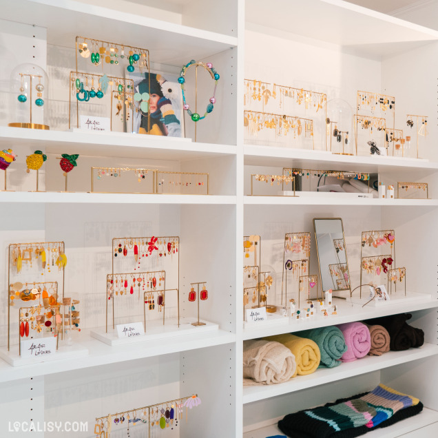 L’intérieur de la boutique "Ariane Lespire" à Liège. Divers bijoux et accessoires colorés sont soigneusement disposés sur des étagères blanches et des présentoirs. Les articles incluent des colliers, des boucles d’oreilles, des bracelets et des bagues, ainsi que quelques textiles pliés.