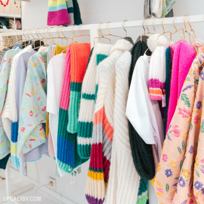 Divers vêtements colorés, notamment des pulls et des écharpes, sont suspendus sur des portants chez "Ariane Lespire" à Liège. Les vêtements présentent une gamme de motifs et de textures, avec des couleurs vives comme le rose, le bleu, le blanc et des imprimés floraux.